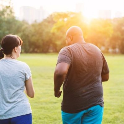 Two people, a man and woman, wear exercise gear outdoors in a green park and walk away from the camera.